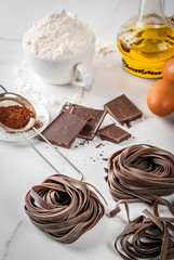 Raw unprepared chocolate pasta noodles, with ingredients for cooking - chocolate, cocoa, flour, eggs,  oil. On a white kitchen marble table. Copy space
