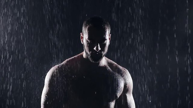 silhouette of a large muscular man bodybuilder in the rain it drains water breaks on a beautiful male body. dark background. slow motion