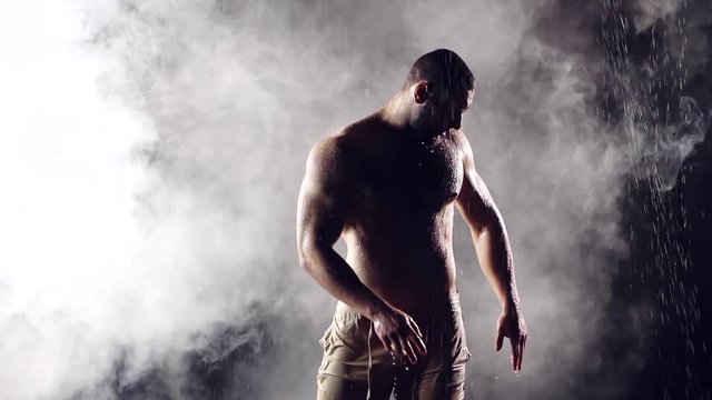 beautiful muscular young male bodybuilder posing under the jets of water against the background of fog