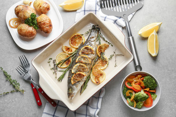 Baking dish with tasty fish, herbs and lemon on grey table