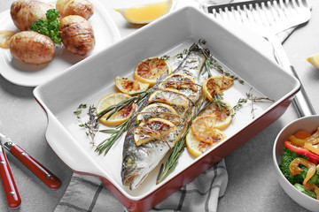 Baking dish with tasty fish, herbs and lemon on grey table