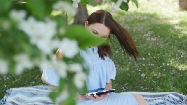 Attractive young woman standing amidst the spring blossom, listening to the music and checking her smartphone. Concept of youth and modern technology