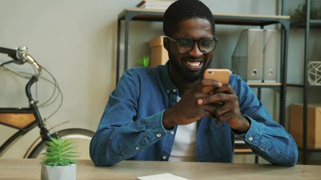 Portrait of smiling african business man in denim blue shirt using smart phone for chating with friends in the work time. Close up.