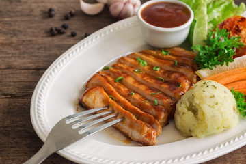 Barbecue pork steak slices on white plate. Delicious pork steak for lunch or dinner on wood table. Moist and soft homemade pork barbecue served with mash potato barbecue sauce and vegetable.