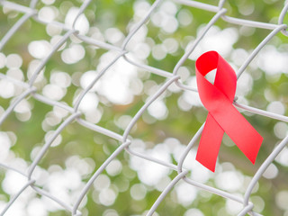 World AIDS day concept. Red ribbon hang on fence mesh netting and tree leaves bokeh background.