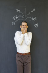 Single white adult man standing in front of a blackboard with drawn social media icons symbolizing frustration, stress and overload from modern communication