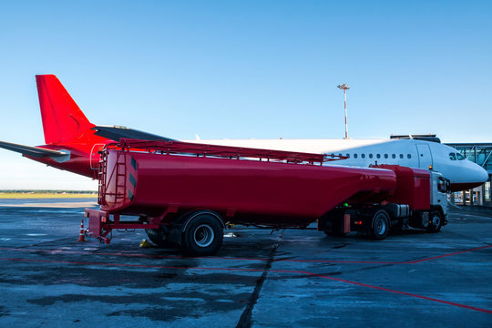 The Red Tanker Refueling The Plane Parked To A Boarding Bridge At The Airport Apron