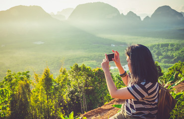 Young traveler woman backpacker use mobile phone take a photo of beautiful of sunrise nature at top of mountain view,Freedom wanderlust concept,Khao Samed Nang Chee,Phang Nga,Thailand