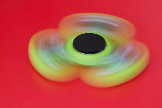 A green hand spinner or fidget spinner is rotating on the red background. A toy for stress relieving. Close up. Selective focus.