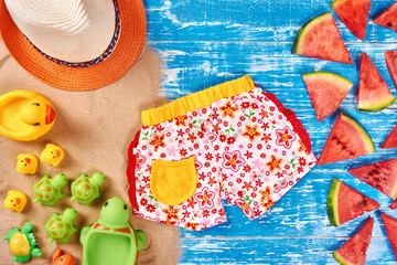 Baby Child summer stuff cloth to the beach on a blue wooden background with sand and watermelon slices. Beach duck and turtles toys. Top view.