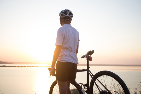 beautiful picture of cyclocross rider in helmet watching sunset or sunrise, lake and hills background 