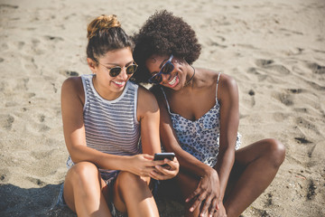 Two female friends on beach using mobile smiling
