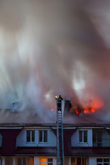 Burning fire flame with smoke on the apartment house roof in the city, firefighter or fireman on the ladder extinguishes fire