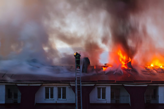 Burning fire flame with smoke on the apartment house roof in the city, firefighter or fireman on the ladder extinguishes fire