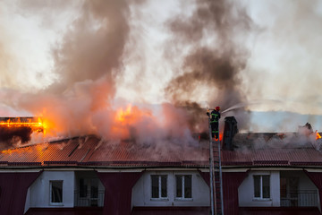 Burning fire flame with smoke on the apartment house roof in the city, firefighter on the ladder extinguishes fire