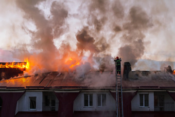 Burning fire flame with smoke on the apartment house roof in the city, firefighter on the ladder...