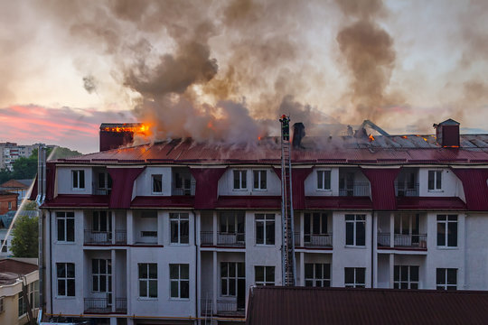 Burning fire flame with smoke on the apartment house roof in the city, firefighter on the ladder extinguishes fire