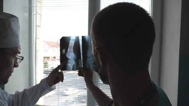 Two doctors view mri picture and discussing about it. Medical workers in hospital examine x-ray prints. Male medics consult with each other while looking at x ray image. Slow motion Rear back view