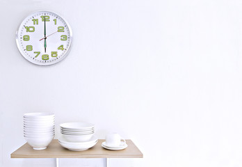 Stack of white bowls, plate and cup on wooden table and white background with clock.