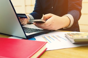 young man holding credit card and using smartphone with report sheet, calculator, laptop on desk, online shopping, online payment, lifestyle technology, finances and economy concept, sunlight effect