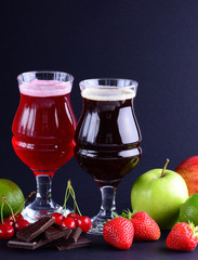 Two wineglasses of craft sweet beer with an assortment of fruits and berries over a black background. Vertical beverage background with a copy space.