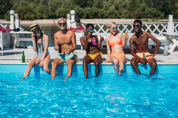 Group of young multiethnic friends sitting with beverages at poolside during vacation at resort