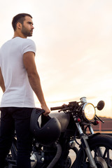 Sporty biker handsome rider man in white t-shirt go travel on classic style cafe racer motorbike on rooftop at sunset. Vintage bike custom made in garage. Brutal urban lifestyle. Outdoor portrait.
