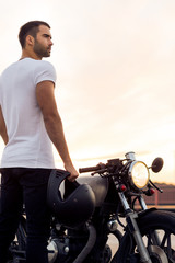 Obraz premium Sporty biker handsome rider man in white t-shirt go travel on classic style cafe racer motorbike on rooftop at sunset. Vintage bike custom made in garage. Brutal urban lifestyle. Outdoor portrait.