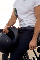 Obraz premium Close up of a handsome rider male in white blank t-shirt with helmet near classic style cafe racer motorcycle at sunset. Bike custom made in vintage garage. Brutal urban lifestyle. Outdoor portrait.