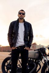 Fototapeta na wymiar Handsome rider man with beard and mustache in black biker jacket, white t-shirt and fashion sunglasses smoking cigaret near classic style cafe racer motorbike at sunset. Brutal fun urban lifestyle.