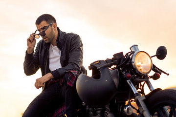 Handsome rider man with beard and mustache in black biker jacket take off sunglasses on classic style cafe racer motorcycle at sunset. Bike custom made in vintage garage. Brutal fun urban lifestyle. - 165690006