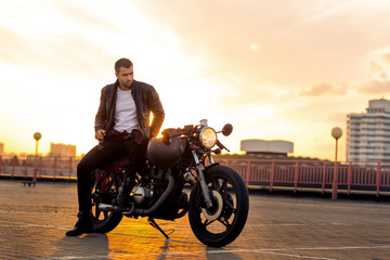 Fototapeta na wymiar Handsome rider guy with beard and mustache in black leather biker jacket sit on classic style cafe racer motorcycle at sunset time. Bike custom made in vintage garage. Brutal fun urban lifestyle.