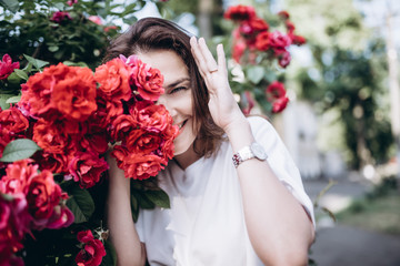 Beautiful portrait of sensual brunette young woman in white dress close to red roses. She is hidding at the rose bush