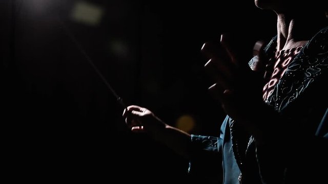 The hands of a female orchestra conductor directing the musicians in the darkness. Medium side shot.

