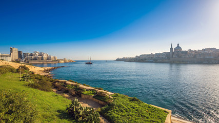 Fototapeta na wymiar Valletta, Malta - Skyline view of the ancient city of Valletta and Sliema at sunrise shot from Manoel island at spring time with sailing boat, St.Paul's cathedral, blue sky and green grass
