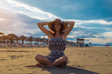 Happy cheerful summertime woman wearing dress and hat sitting on the beach and holding hat with sunshades and blue sky and sea in the background