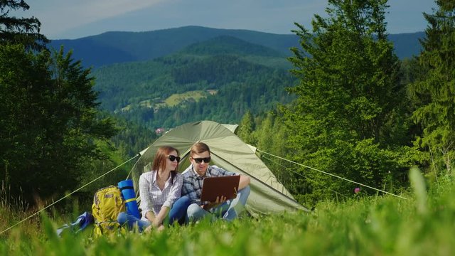 A young couple is resting in a camping near a tent, enjoying a laptop. Technology in nature