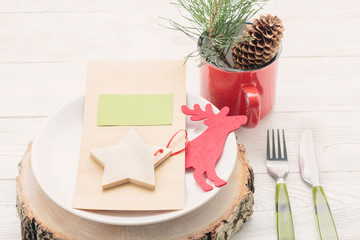 Christmas dinner laying concept. White plate on wooden board with menu card, fork, knife, red mug with Christmas fir twig decor, deer figure, wooden star with green label on white wooden table