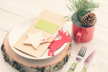 Close-up of Christmas dinner laying. Plate on wooden board with menu card, fork, knife, red mug with Christmas decor, deer figure, wooden star with green label. Christmas restaurant laying decoration