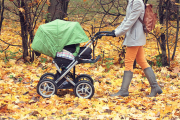 Fototapeta premium Mother pushing stroller in autumn park. Young woman strolling with a baby in buggy outdoors. Dry orange leaves on the ground