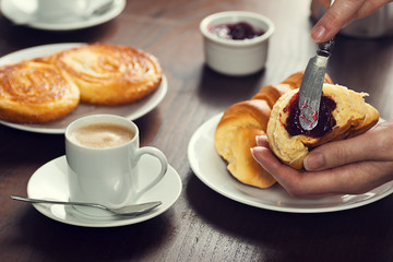 Woman's Hand Spreading Fruit Preserves (Jam) Across a Croissant Bread Roll with a Knife in a Cafe