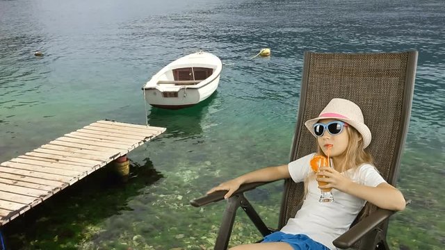 Young caucasian girl sitting on armchair and drinking orange juice cocktail through straw. Serious young girl looking. Lonely boat swaing on waves of sea. Wooden pier at green water sea.