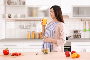 Young pregnant woman drinking juice while standing near table in kitchen