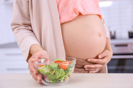 Young pregnant woman near table with bowl of salad in kitchen