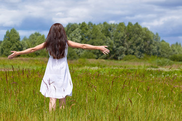 Naklejka na ściany i meble Beauty girl enjoying nature. Beautiful teenage model girl in white dress raised hands up on the spring field. Freedom and religion concept. Green grass and blue clouds.