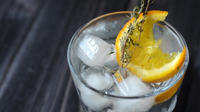 Close-up of a coctail in a glass with ice, a piece of orange and a sprig of rosemary. Macro view and zoom camera on the object. Cocktail with citrus fruits on dark wooden table