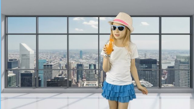 Young girl walks in empty office and drinks orange juice. Behind panoramic windows can see cityscape and skyscrapers. Girl quenches her thirst with an orange cocktail. Good health.
