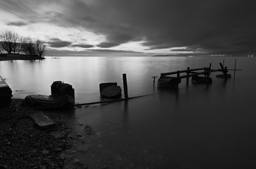 A pier built of wood and stones on a lake at dusk, with perfectly still water and moving clouds