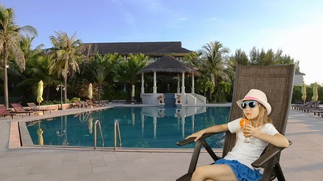 Young caucasian girl sitting on armchair and drinking orange juice cocktail through straw. Girl sitting near sweaming pool with blue water. Have same clip with transparent background Alpha Channel.