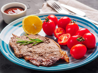 Close up view of fried steak served with fried potatoe sherry tomatoes in blue plate on black concrete background